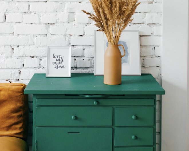 image of a green dresser, with 2 paintings and a beige plant pot and a white background wall with brick wallpaper